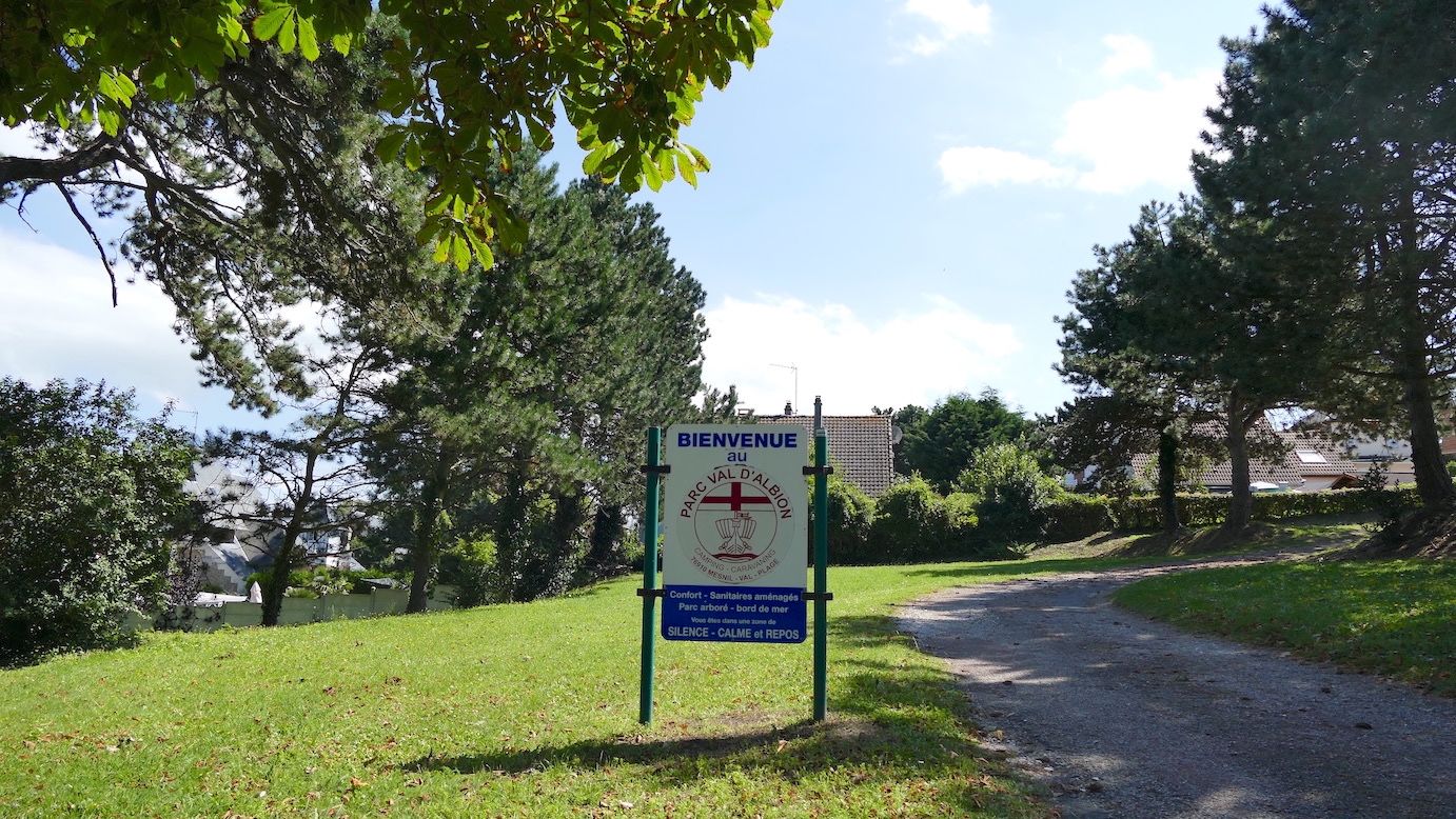Schild mit dem Campingplatznamen auf einer Wiese neben einem kleinen Weg.