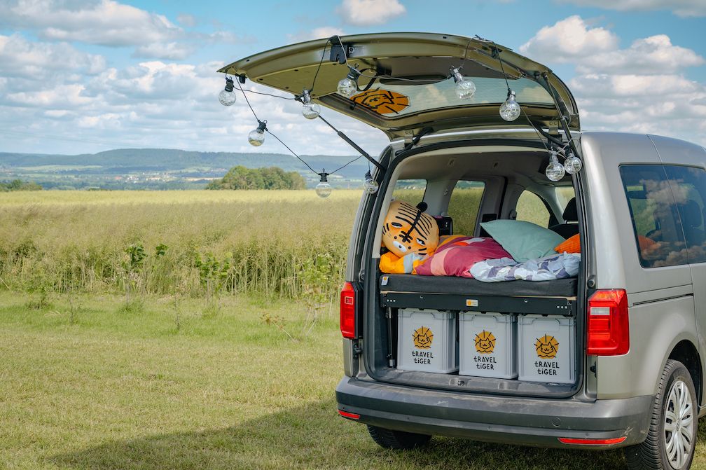 Caddy steht mit offener Heckklappe und aufgebautem Campingbett auf Wiese.