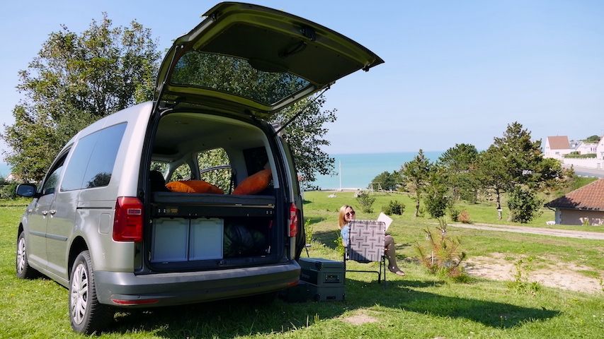 Frau sitzt lesend neben ihrem Minicamper mit Blick aufs Meer.