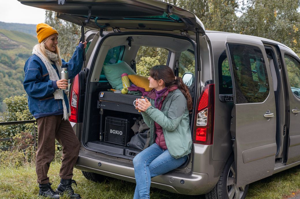 Zwei Mädchen trinken Tee am Kofferraum eines Minicampers.