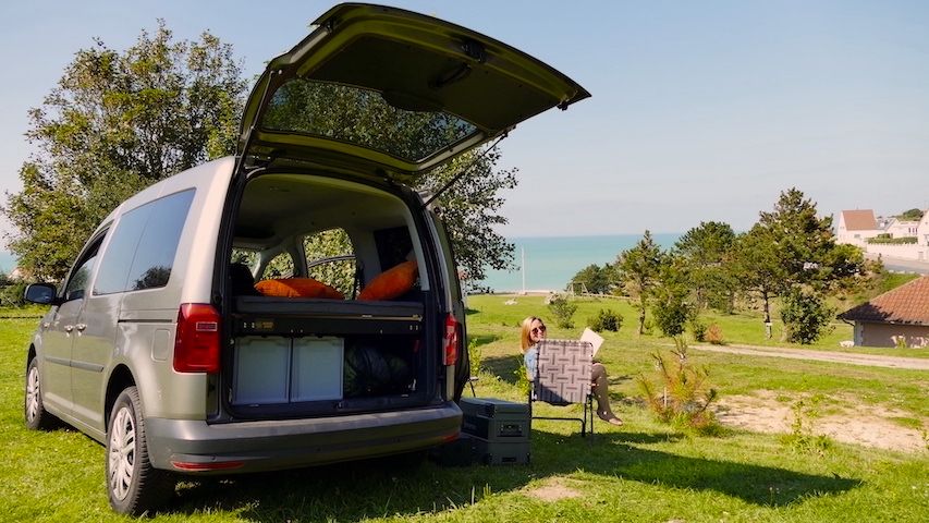 Anna sitzt neben dem Caddy Camper auf einem Stellplatz mit Meerblick im Campingstuhl und liest. 