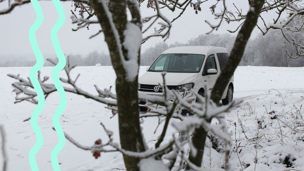 Ein VW Caddy steht auf einer verschneiten Wiese am Waldrand. 