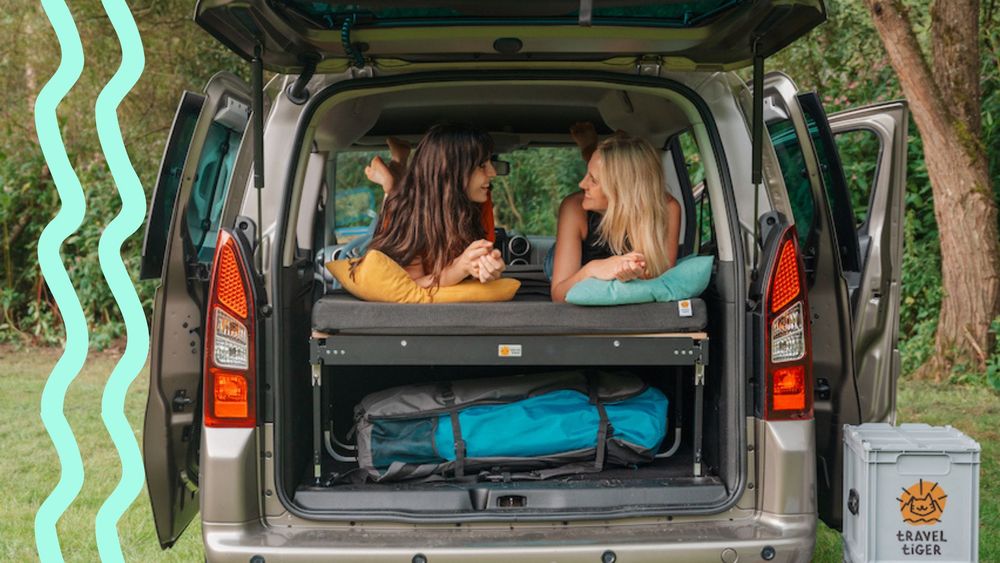 Zwei junge Frauen liegen lachend auf einem Campingbett im Auto.