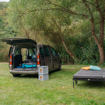 Ein Minicamper steht mit einem freistehenden Campingbett auf einer Wiese.